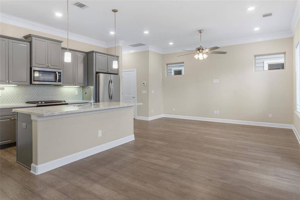This open-concept space features a modern kitchen with gray cabinetry, stainless steel appliances, and a large island with pendant lighting. It flows into a spacious living area with wood flooring, a ceiling fan, and ample natural light from the windows. Ideal for entertaining and family gatherings.