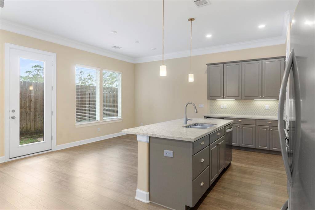 This open-concept space features a modern kitchen with gray cabinetry, stainless steel appliances, and a large island with pendant lighting. It flows into a spacious living area with wood flooring, a ceiling fan, and ample natural light from the windows. Ideal for entertaining and family gatherings.