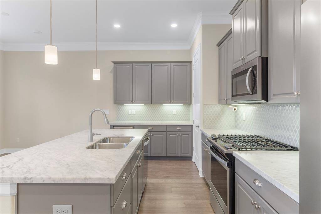 This open-concept space features a modern kitchen with gray cabinetry, stainless steel appliances, and a large island with pendant lighting. It flows into a spacious living area with wood flooring, a ceiling fan, and ample natural light from the windows. Ideal for entertaining and family gatherings.