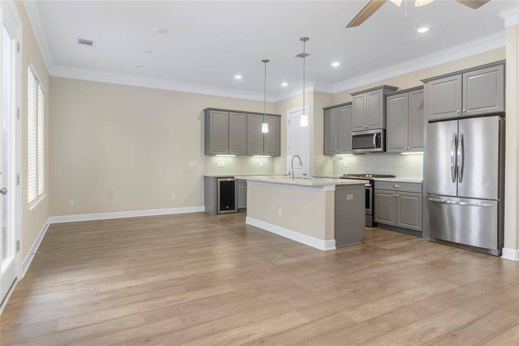 This open-concept space features a modern kitchen with gray cabinetry, stainless steel appliances, and a large island with pendant lighting. It flows into a spacious living area with wood flooring, a ceiling fan, and ample natural light from the windows. Ideal for entertaining and family gatherings.