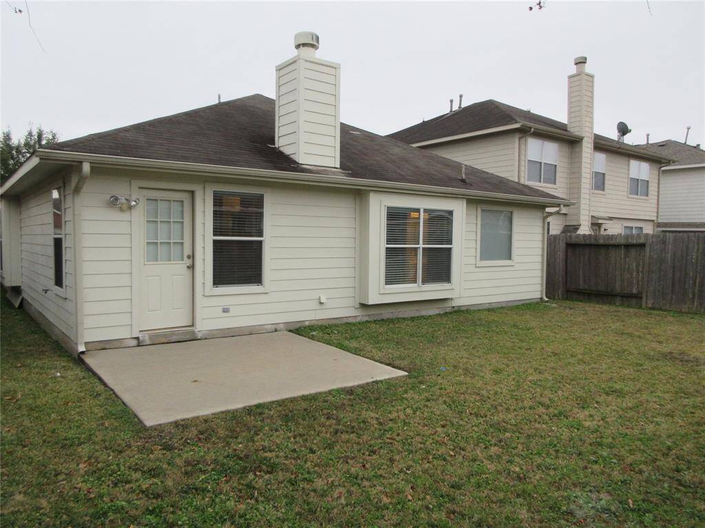 The house features the backyard of a single-story house with a small, paved patio area. It has light-painted siding, a chimney, and several windows. The yard is grassy and enclosed by a wooden fence, offering a private outdoor space.
