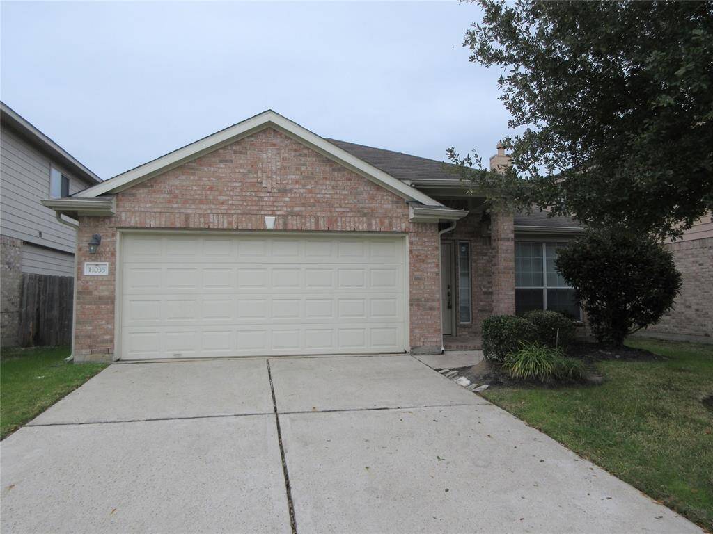 This beautiful single-story brick house with a two-car garage, features a simple front yard with a paved driveway and some shrubs next to the entrance. The roof is gabled, and there's a window to the right of the entrance.