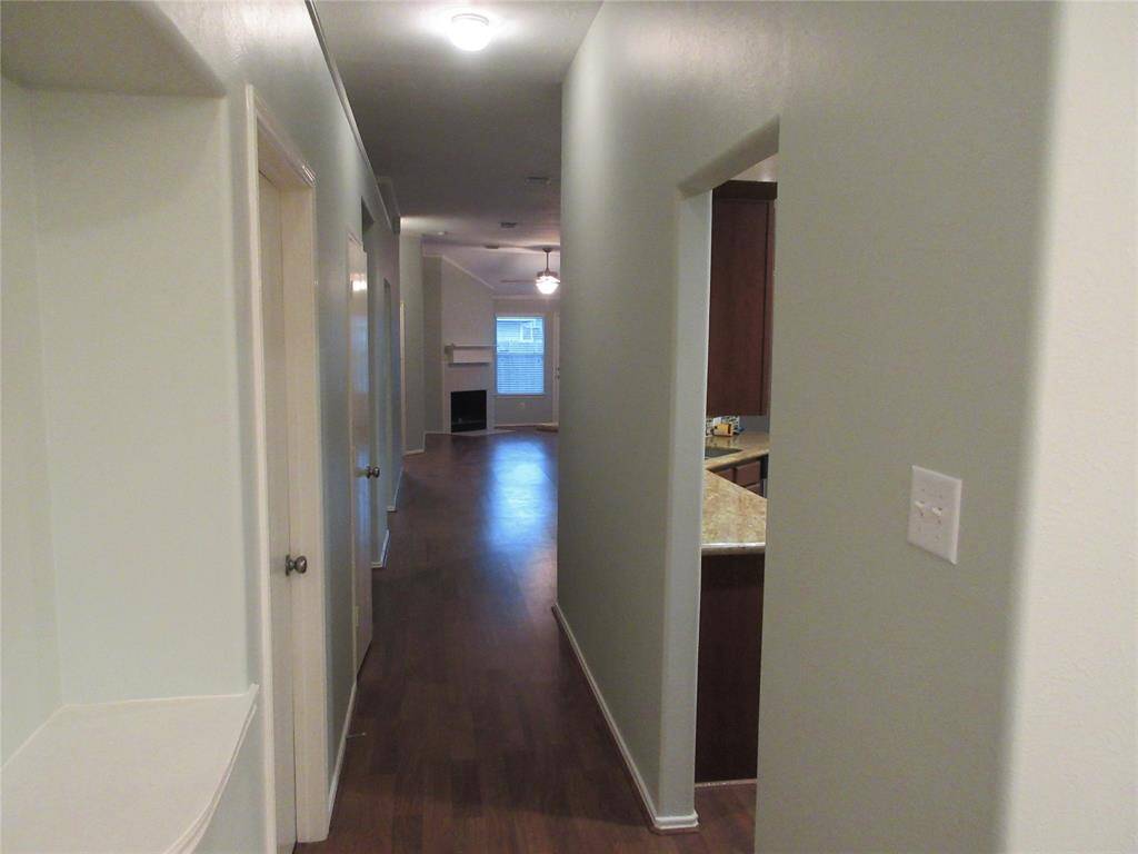 The hallway leads to a living area with a fireplace and large window. The hallway connects several rooms, and there's a glimpse of a kitchen with granite countertops on the right.