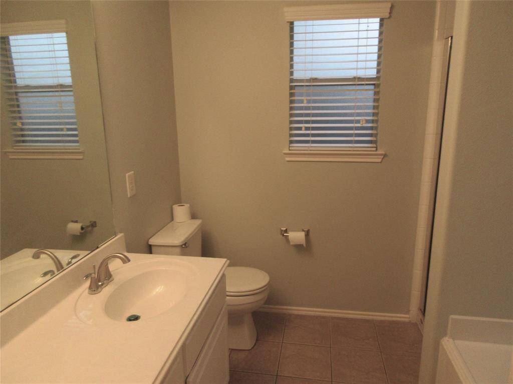This bathroom features a clean, neutral design with a white vanity, sink, and toilet. It has tiled flooring, a large mirror, and two windows with blinds, providing good natural light.