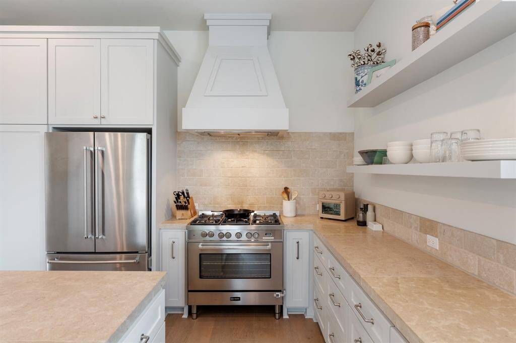 A closer look at the kitchen’s cooking area reveals a premium 6-burner gas range framed by custom vent hood and a textured tile backsplash. Floating shelves adds a stylish touch and functional display.