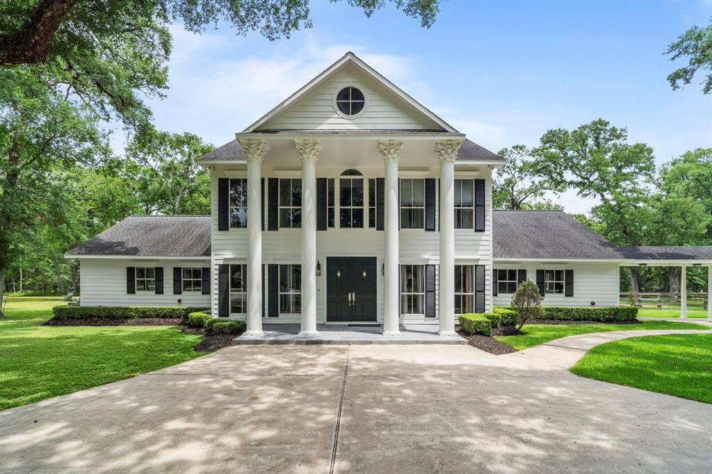 Elegant two-story Colonial-style home with grand columns and symmetrical design. Features a spacious driveway, manicured lawn, and mature trees, offering a serene and upscale setting.