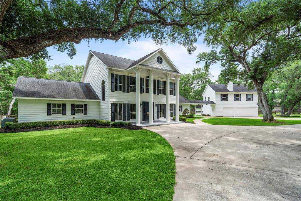 A classic two-story home with a grand front porch supported by columns. The property is surrounded by lush greenery and mature trees, featuring a spacious driveway and a detached garage.