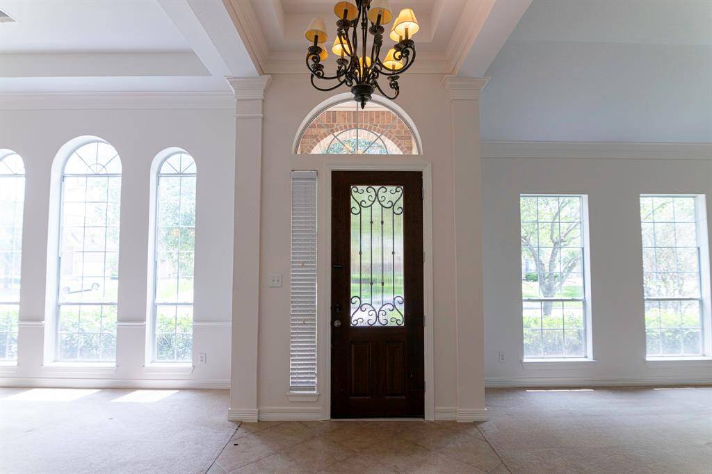 This entryway features a grand wooden door with ornate ironwork, surrounded by large arched and rectangular windows that allow plenty of natural light. The space is accentuated by a stylish chandelier and high ceilings, creating an inviting and airy atmosphere.
