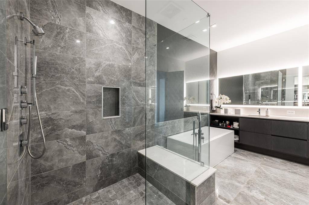 A thoughtfully designed primary bathroom featuring porcelain tile extending to the ceiling, accented by a striking designer wallpaper wall, custom lighting around the mirror, and LED lighting throughout.