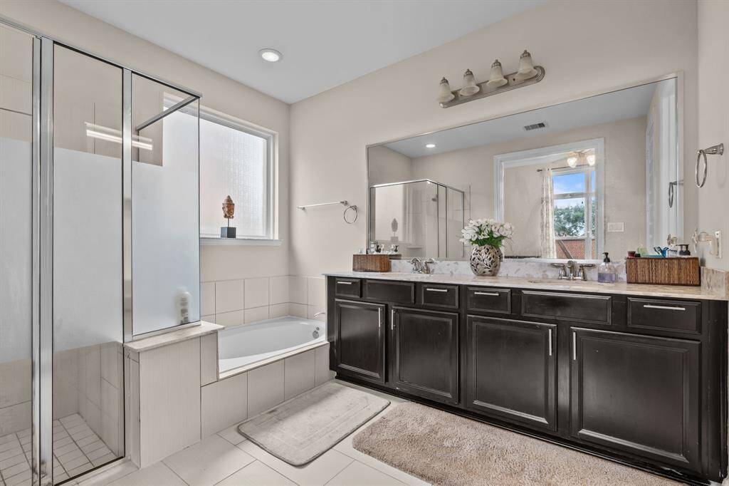 This spacious bathroom features a glass-enclosed shower, a separate bathtub, and a double vanity with dark cabinetry. The large mirror enhances the natural light from the window, creating a bright and inviting space.