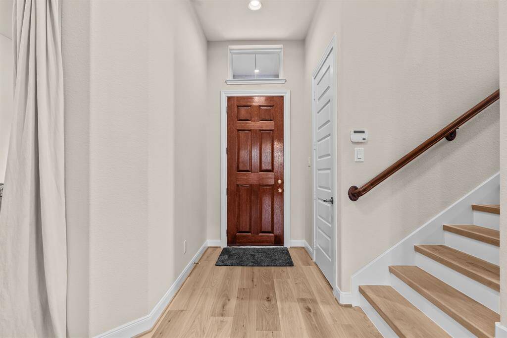 A welcoming entryway features a wooden front door with a transom window, light wood flooring, and a staircase with wooden handrails. Neutral walls create a bright, open atmosphere, complemented by a side door and modern thermostat.