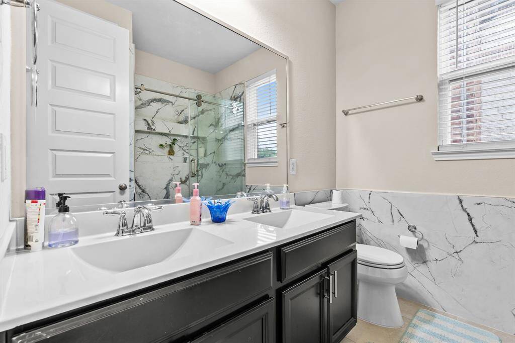 Bright and modern bathroom featuring a double vanity with sleek faucets, large mirror, and a spacious glass shower with elegant marble-style tiles. Natural light streams through a window, highlighting the clean, contemporary design.