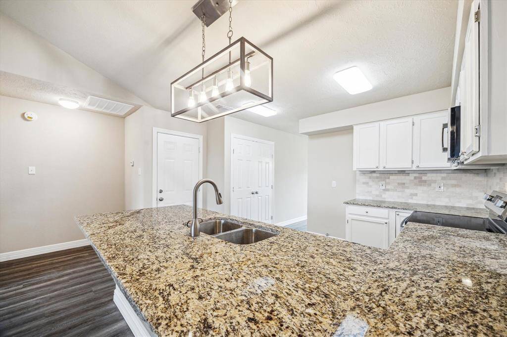Modern kitchen with a spacious granite countertop, double sink, and stylish pendant lighting. Features white cabinetry, tile backsplash, and brand new appliances, complemented by a neutral color palette.