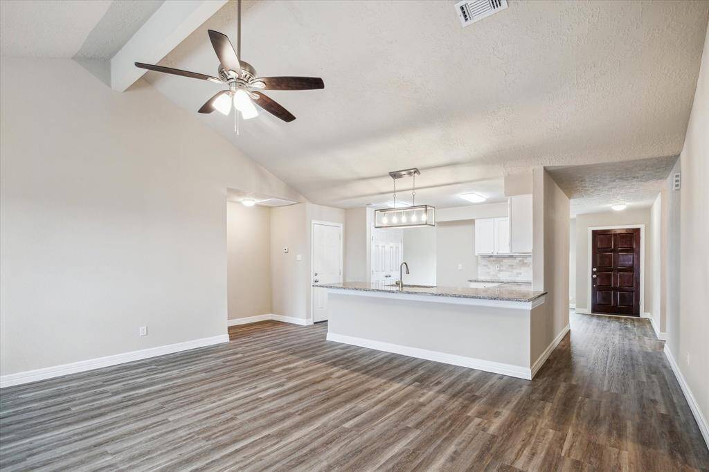 This photo showcases a bright, open-concept living area with modern wood-look flooring, a ceiling fan, and a stylish kitchen with white cabinetry and granite countertops. The space is well-lit and features neutral tones, offering a welcoming atmosphere.