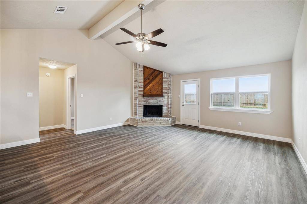 This spacious living room features a vaulted ceiling with a fan, a charming brick fireplace, and large windows for natural light. The room has stylish wood like flooring and neutral walls, creating a bright and inviting atmosphere.