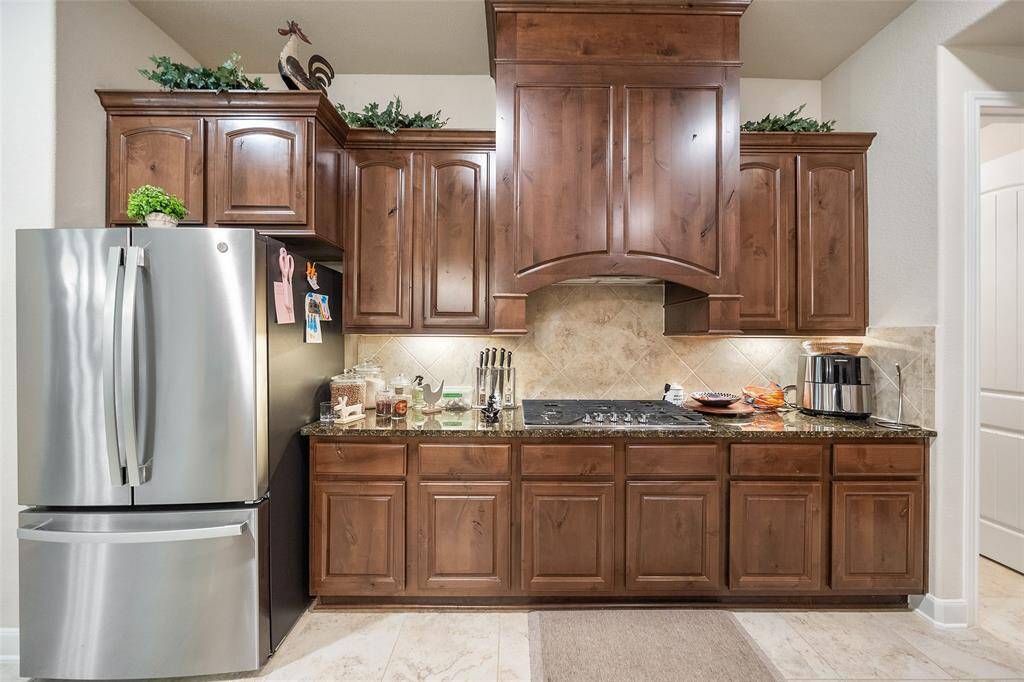 The built-in gas cooktop with an impressive wood hood vent is ready for weeknight dinners or weekend feasts, set against rich wood cabinetry, granite countertops, under-cabinet lighting, and tile backsplash. It’s a warm, functional space designed for real-life cooking.
