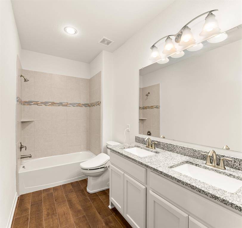 This bathroom features a modern design with a double vanity, granite countertops, and stylish lighting. It includes a bathtub with elegant tile work and wood-look flooring, combining functionality with contemporary aesthetics