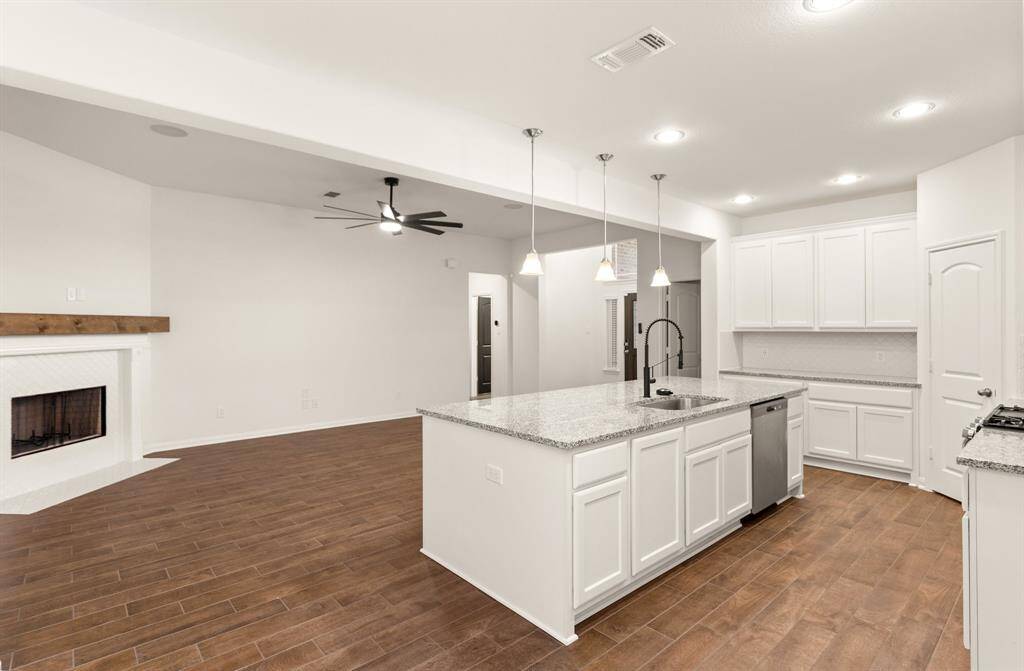 This spacious kitchen and living area features an open floor plan with sleek wood flooring, a modern fireplace, and a large island with granite countertops. The kitchen includes stainless steel appliances, ample cabinet space, and stylish pendant lighting