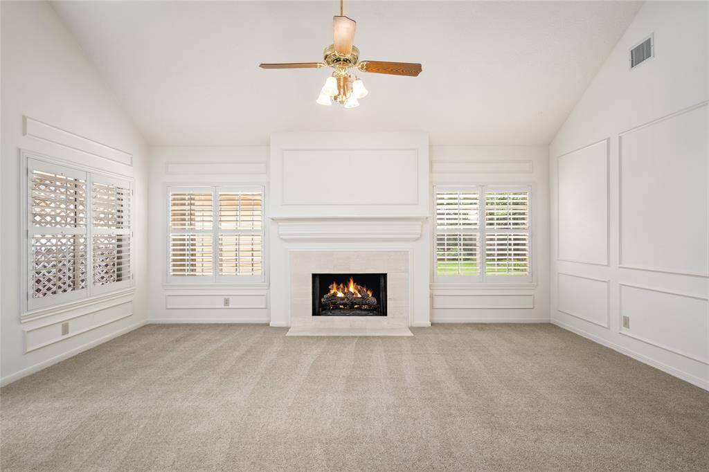 Spacious living room features a cozy gas fireplace as the focal point, complemented by elegant wall molding and a soaring vaulted ceiling. Natural light pours in through large windows with classic plantation shutters, creating a bright and inviting atmosphere. Neutral carpet adds warmth and comfort to the space.