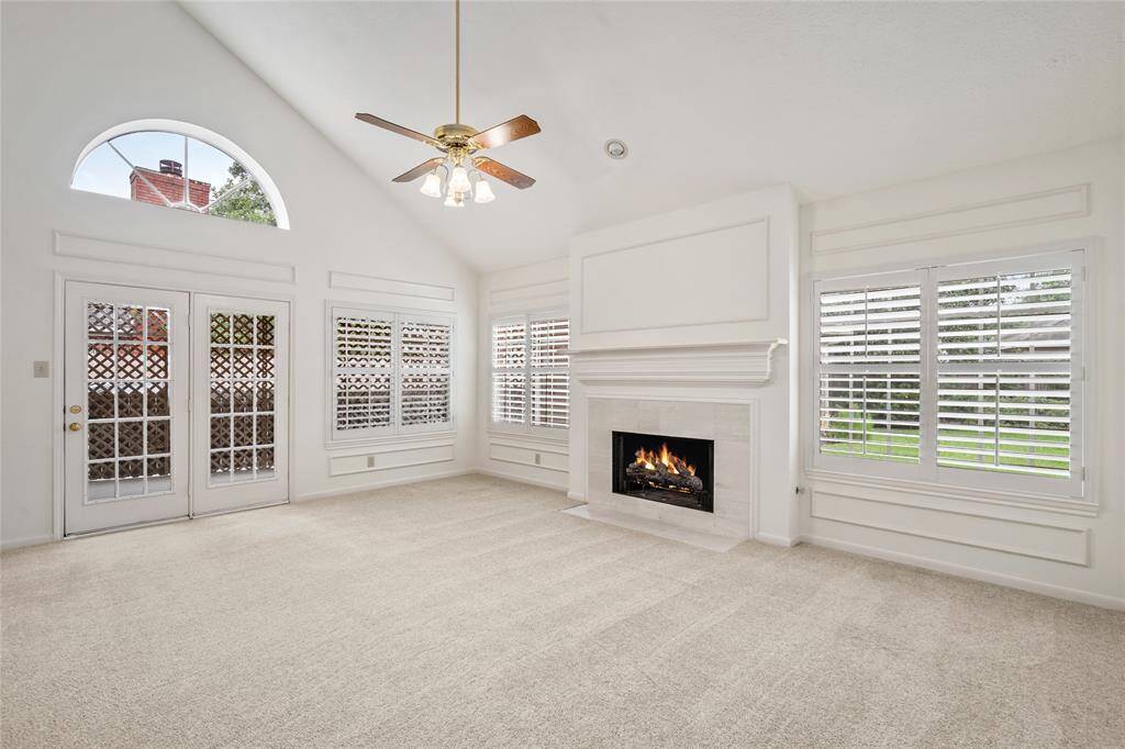 Elegant living area showcases a stately gas fireplace with a detailed mantel, framed by crisp white panel molding and vaulted ceilings. Abundant natural light flows through plantation-shuttered windows, offering serene views and timeless charm. A soft neutral palette and plush carpeting create a warm, welcoming setting