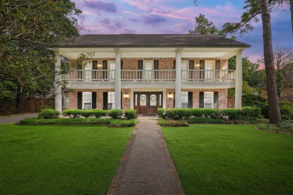 Classic red brick exterior with tall columns, black shutters, and an inviting second-story balcony. A long pebble and brick walkway leads through a meticulously maintained lawn to stunning double front doors—an elegant welcome to this Georgia-style charming home.