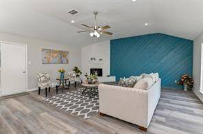 This spacious living room features modern laminate flooring, a bold blue accent wall, and a cozy fireplace. It has a ceiling fan, recessed lighting, and ample natural light through large windows. The room is tastefully furnished with a comfortable sofa and stylish chairs, making it ideal for relaxing or entertaining.