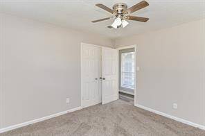 A bright, neutral-toned bedroom with carpeted flooring features a ceiling fan and an open doorway leading to a well-lit hallway.