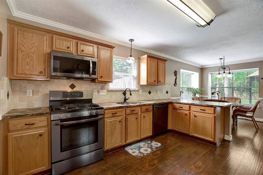 This kitchen features wood cabinetry, modern black stainless steel appliances, and granite countertops. It opens to a breakfast bar & breakfast area with large windows, offering a view of the backyard.