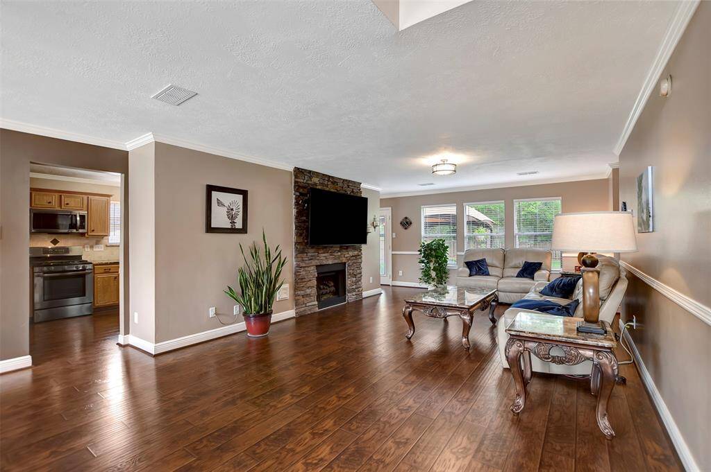 The living room features a stone fireplace with a mounted TV, and natural light from large windows. It's connected to an open kitchen area, offering an inviting atmosphere for gatherings.