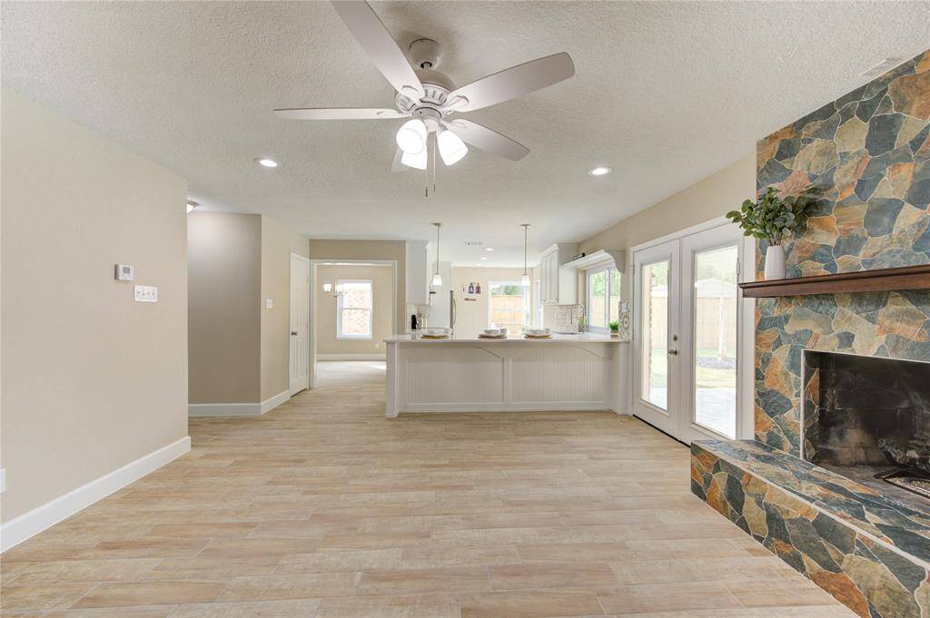 This open-concept living area features a modern kitchen with a breakfast bar, a cozy stone fireplace, and ample natural light from large windows and glass doors leading to the backyard. Its neutral color palette and wood-like tile flooring, creating a warm and inviting atmosphere.
