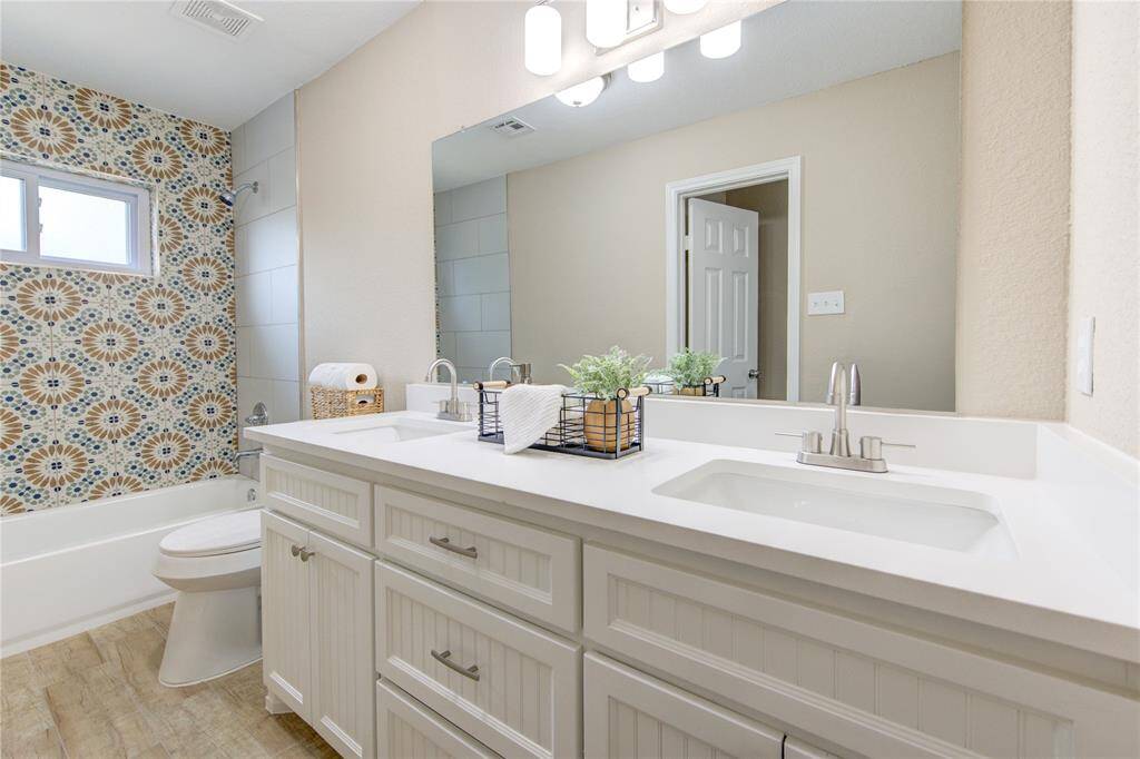 Check out this huge guest bath! This bathroom features a double vanity with modern faucets, a large mirror, custom built cabinetry with ample storage. The tub-shower combo boasts a striking geometric tile design, complemented by natural-toned flooring.