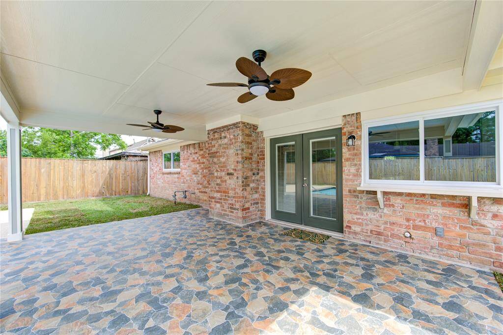 This photo showcases a spacious covered patio with a tiled slate floor, ceiling fans, and brick exterior walls. It features double glass doors leading inside and overlooks a fenced backyard, ideal for outdoor relaxation and entertainment.
