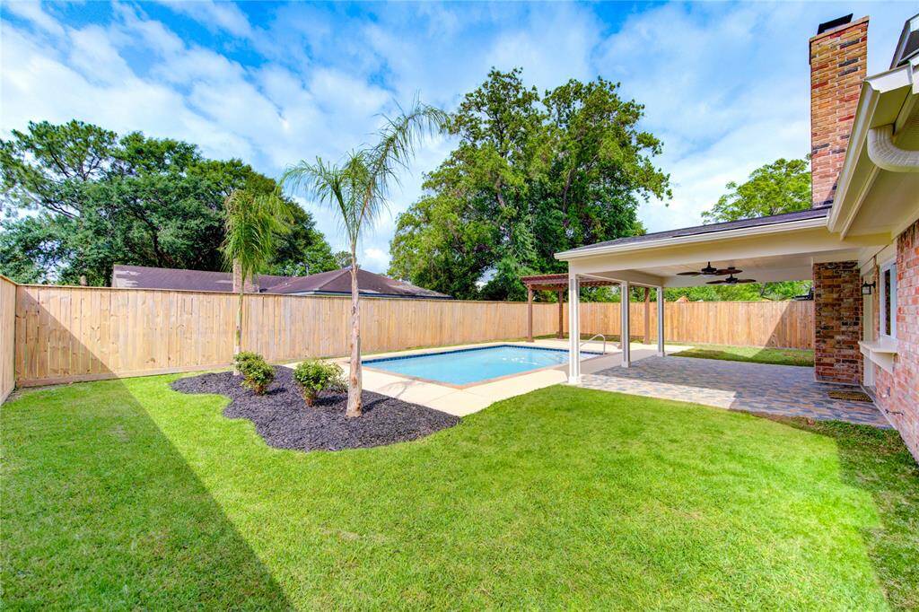 Charming backyard featuring a pool, covered patio with ceiling fans, and landscaped garden with palm trees, all enclosed by a privacy fence. Ready for relaxing and entertaining.