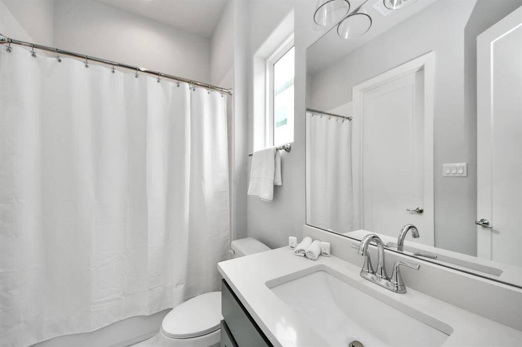 This bathroom features a clean, modern design with a white vanity and chrome fixtures. It includes a bathtub with a shower curtain, a large mirror, and a window for natural light, offering a bright and fresh feel.