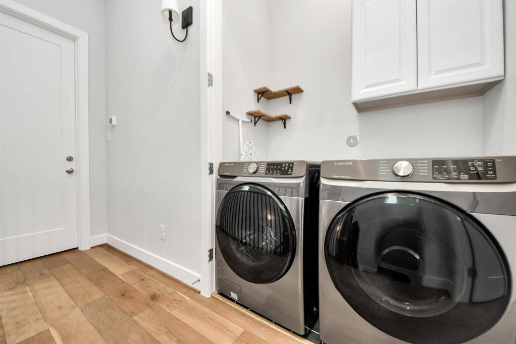 This photo shows a modern laundry room featuring sleek Samsung washer and dryer units, hardwood flooring, and wall-mounted storage with shelves and cabinets.