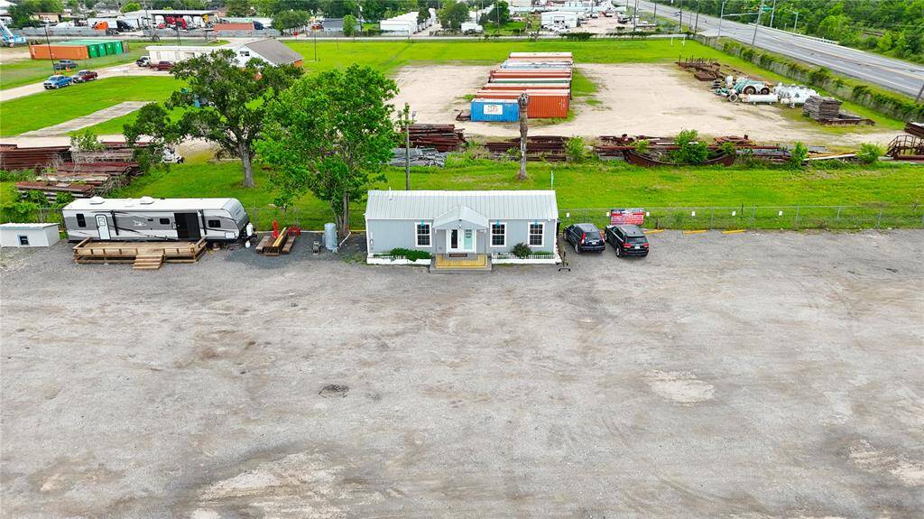 This photo shows a small, single-story home with a metal roof, situated on a spacious gravel lot. There are two cars parked in front and a large RV parked to the left. The property is bordered by industrial storage containers and open green space. It's located near a highway, providing easy access to nearby amenities.