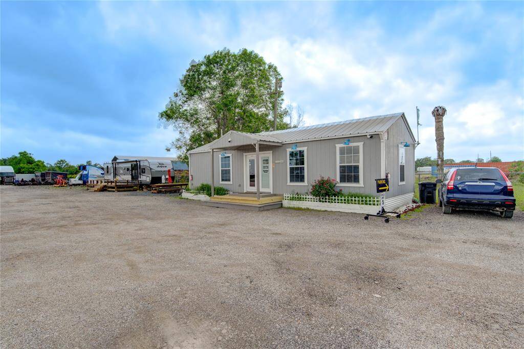This photo shows a small, gray modular home with a metal roof situated in a spacious gravel lot. There are a few parked vehicles, including a car and trailers, and some greenery around the house. The setting has an open, rustic feel with a clear blue sky overhead.