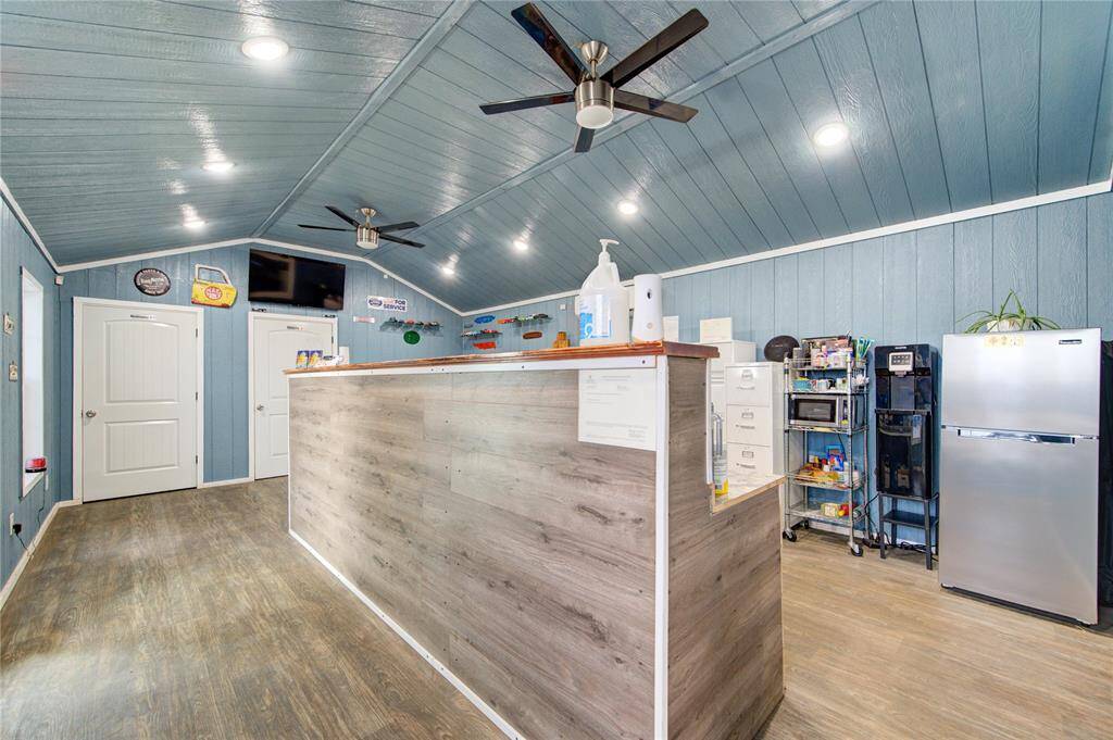 This photo showcases a spacious, modern room with a high, angled ceiling and wood-paneled walls painted in a soft blue. It features a long wooden counter, ceiling fans, recessed lighting, and has practical amenities like a fridge and coffee station, making it ideal for a multi-purpose space.
