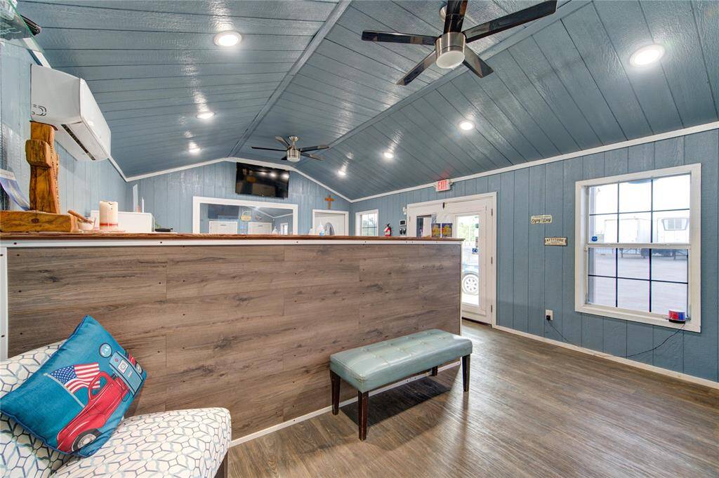 This photo showcases a modern, spacious room with blue paneled walls and ceiling, featuring recessed lighting and ceiling fans. It includes a wooden counter, a comfortable bench, and large windows that let in natural light. The room has a welcoming and functional design, ideal for a reception or lounge area.