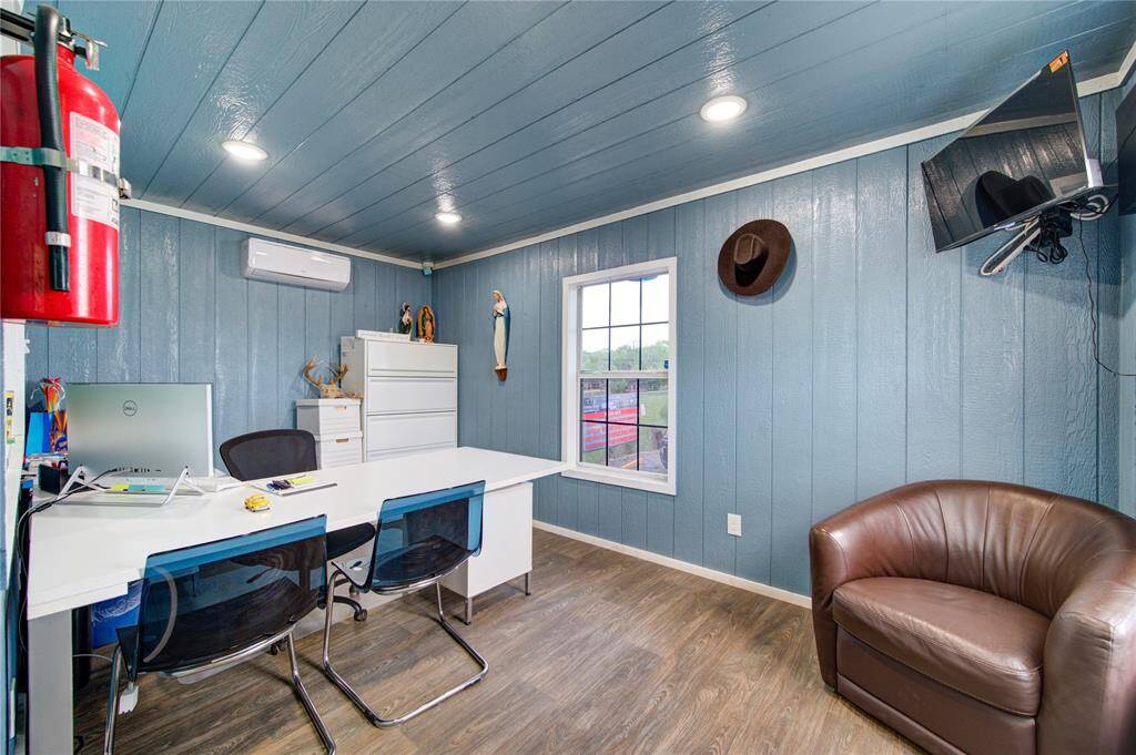 This photo shows a cozy, well-lit office space with blue paneled walls and wood flooring. It features a desk with chairs, a filing cabinet, wall-mounted TV, and a window with a view. A leather chair and decorative items add to the room's appeal.