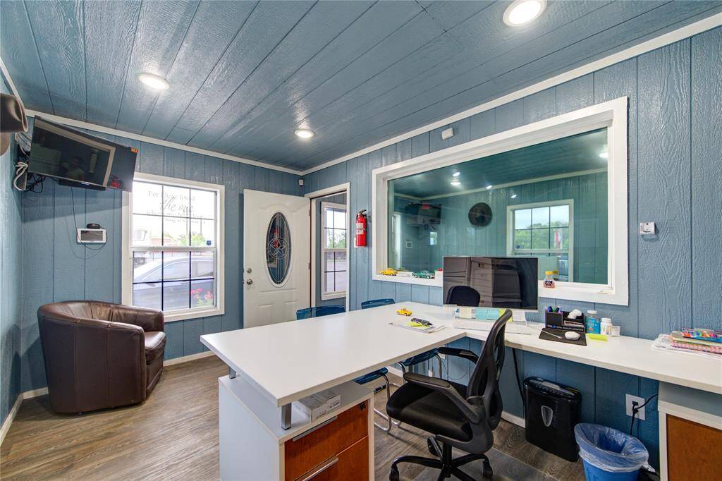 This photo shows a cozy office space with blue paneled walls and wood flooring. It features a white desk, a comfortable chair, and a large window, allowing for natural light. There's a mounted TV and a leather chair, creating a welcoming and functional work environment.