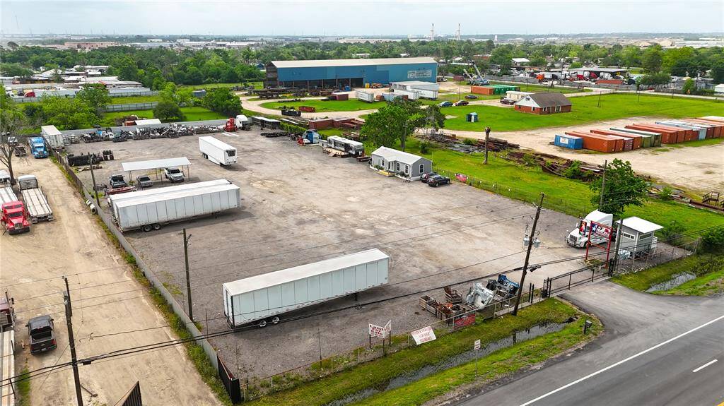 This photo shows a commercial property with a large gravel lot, ideal for industrial use or parking. It includes trailers and equipment, surrounded by fencing, and is situated in a semi-industrial area with nearby green spaces and various commercial buildings.