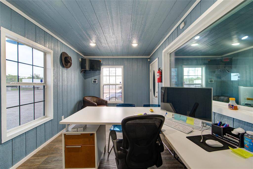 This photo showcases a compact, well-lit office space with blue wood-paneled walls and modern amenities. It features a large desk, ergonomic chair, wall-mounted TV, and ample natural light from two windows. Perfect for a functional and cozy workspace.
