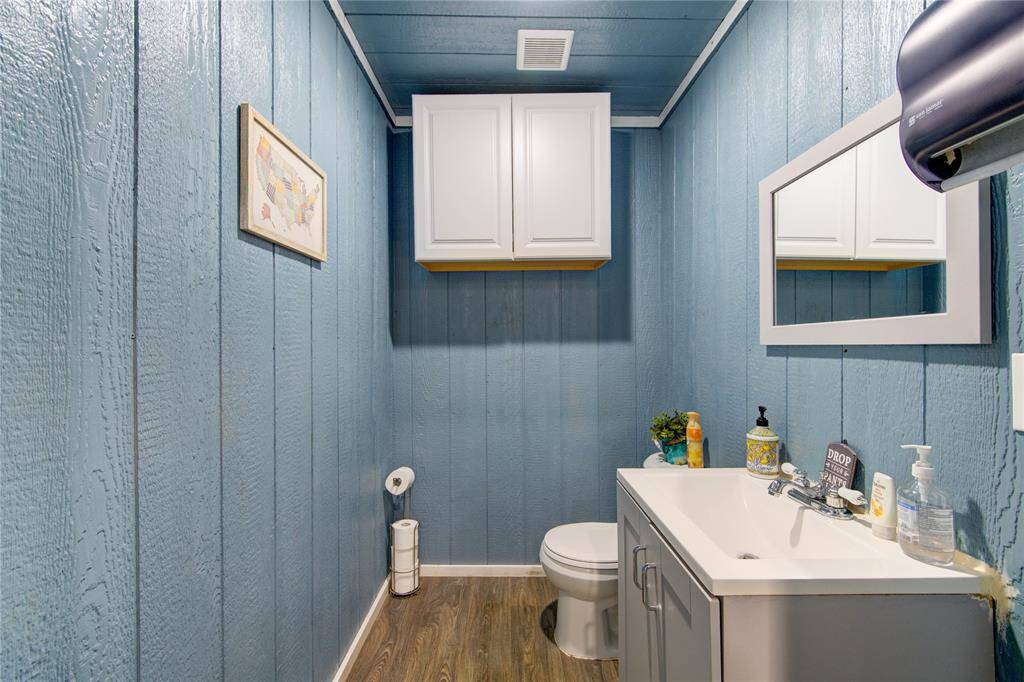 This bathroom features rustic blue wood-paneled walls, a modern white vanity with a mirror, and overhead storage cabinets. The space is accented with wood-look flooring and simple decor for a cozy feel.