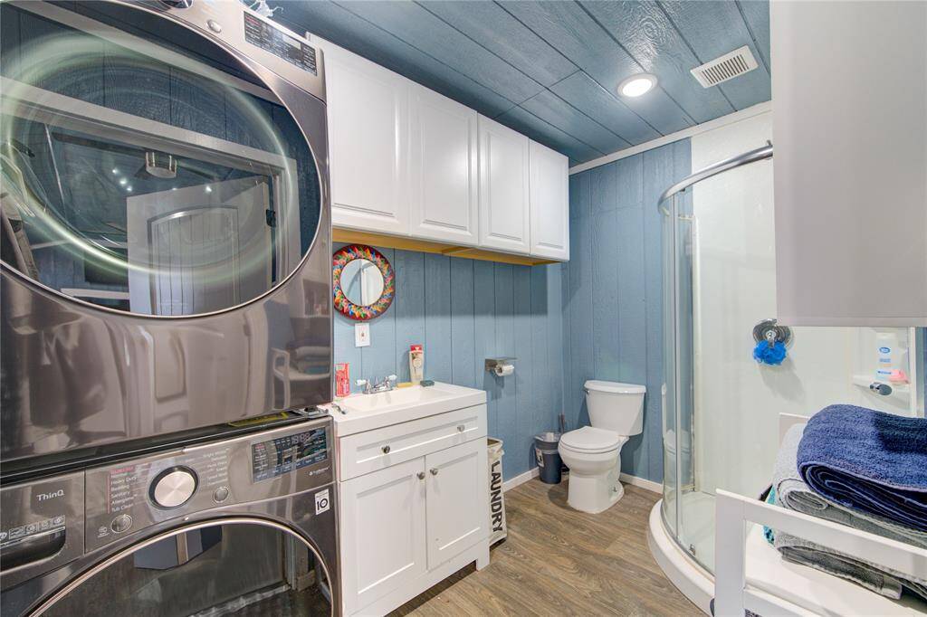 This photo shows a modern bathroom with a compact layout, featuring a stacked washer and dryer, a glass-enclosed corner shower, and a toilet. The room has a blue accent wall, a white vanity with storage, and wood-look flooring, creating a functional and stylish space.
