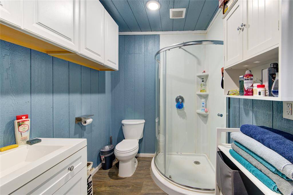 This bathroom features a compact, modern design with light blue wood-paneled walls and a matching ceiling. It includes a curved glass shower, white cabinetry for ample storage, and a sleek white sink. The space is efficiently organized and stylishly appointed.