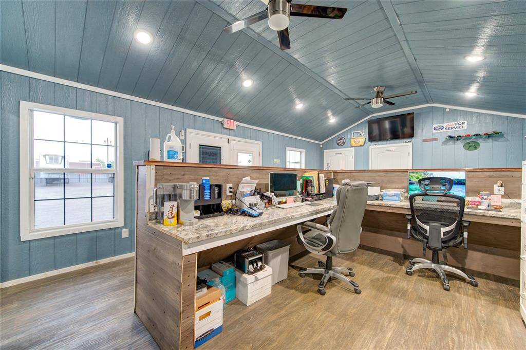This photo shows a spacious, well-lit office with a vaulted ceiling, featuring blue paneling and wood floors. The room includes a large desk with multiple workstations, modern chairs, and ceiling fans, creating a comfortable and efficient workspace.