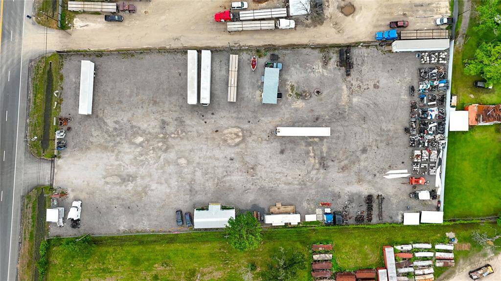 This aerial photo shows a large, gravel-paved commercial lot with various vehicles and equipment. It's bordered by a road on one side and has some greenery around the perimeter, along with a few small structures. Ideal for industrial use or storage.