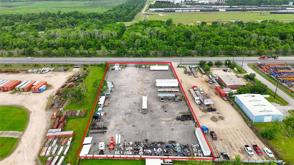 This aerial photo shows a large, fenced industrial lot with several structures and parked vehicles. It's bordered by greenery and adjacent to a major road, offering ample space for commercial use.