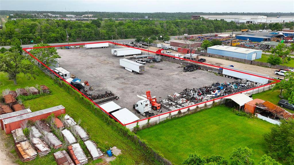 This photo shows a large, fenced industrial lot with a paved surface, suitable for commercial use. It contains several trucks and equipment, and is surrounded by greenery and other industrial buildings.